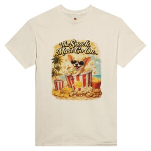 Könnte beinhalten: Cremefarbenes T-Shirt mit einer Cartoon-Illustration eines Chihuahuas mit Sonnenbrille in einem Popcorn-Eimer. Das Bild enthält Snacks, ein Getränk und den Text "The Snack Must Go On."