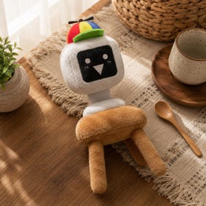 May include: A plush toy featuring a white, square-shaped head with a black screen face displaying two white eyes and a triangular nose. The toy wears a colorful propeller cap with red, yellow, blue, and green panels. It has a brown, rounded body.