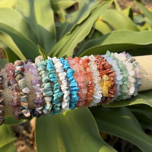 May include: A collection of gemstone chip bracelets in various colors, including brown, purple, turquoise, red, and green. The bracelets are displayed on a beige cylindrical stand, set against a backdrop of green leaves.