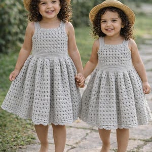May include: Two young children wearing matching gray crochet dresses and straw hats. The dresses have a square neckline and a full skirt. They are holding hands and smiling, walking on a stone path.