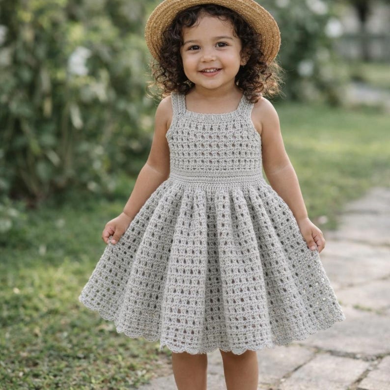 May include: A light gray crochet dress with a flared skirt and square neckline. The dress has a scalloped hem and is worn by a young child. The child is also wearing a straw hat and smiling.