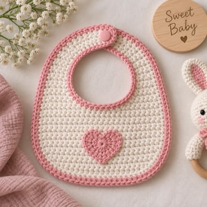 Pode incluir: Um babador de crochê para bebê na cor creme com uma aplicação de coração rosa no centro e uma borda rosa. O babador possui um fecho de botão de pressão rosa na parte superior. O design é simples com um padrão de pontos texturizado.