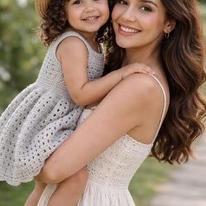 May include: A woman holding a young child. The child wears a straw hat and a light gray crochet dress. The woman wears a white eyelet dress. Both are smiling. The background is blurred green foliage.