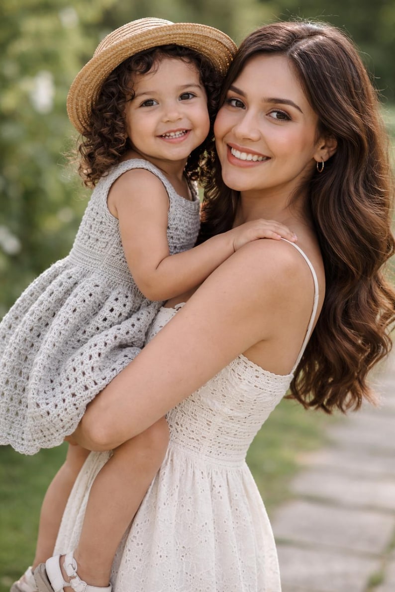 May include: A woman holding a young child. The child wears a straw hat and a light gray crochet dress. The woman wears a white eyelet dress. Both are smiling. The background is blurred green foliage.