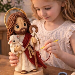 May include: A handcrafted crochet figure of Jesus holding a lamb, with a halo and staff. The figure is dressed in cream and burgundy robes, with brown hair and beard. The figure is on a wooden base.