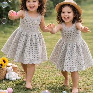 May include: Two young children wearing light gray crochet dresses and straw hats play outdoors. They are smiling and reaching for bubbles. The dresses have a scalloped hemline. A basket with sunflowers and a stuffed bear are in the background.