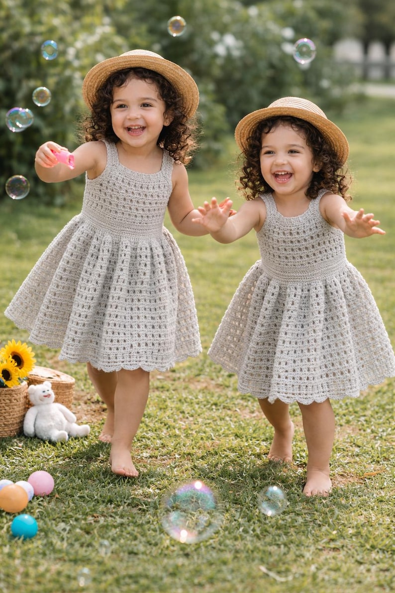 May include: Two young children wearing light gray crochet dresses and straw hats play outdoors. They are smiling and reaching for bubbles. The dresses have a scalloped hemline. A basket with sunflowers and a stuffed bear are in the background.