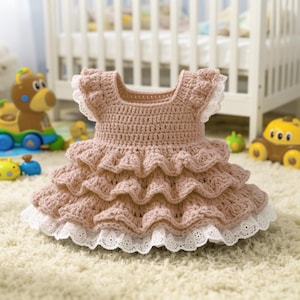 May include: A light pink, handmade crochet dress for a baby or toddler. The dress has a square neckline, ruffled sleeves, and a tiered skirt with white lace trim. The dress is displayed on a white carpet, with toys visible in the background.