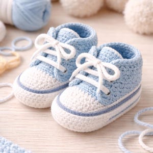 May include: A pair of baby blue and white crochet sneakers with white laces. The shoes have a blue trim and are made of soft yarn. The sneakers are designed for infants and are displayed on a light wooden surface.