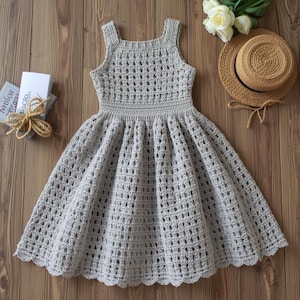 May include: A light gray, handmade crochet dress with a square neckline and a flared skirt. The dress features a textured open weave design. A small straw hat and white flowers are next to the dress.