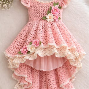 Peut inclure: Une robe rose en crochet faite à la main, avec une jupe à volants et des ornements floraux. La robe présente un col rond, des manches courtes et un ourlet asymétrique. Des fleurs roses et blanches sont attachées au corsage et à la jupe, ajoutant une touche d'élégance.
