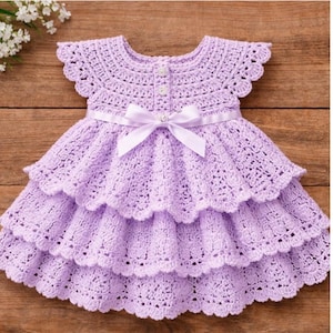 May include: A lilac crochet dress with a tiered skirt and a ribbon bow at the waist. The dress has a scalloped neckline and sleeves, and is buttoned down the front. The dress is designed for a baby or toddler.