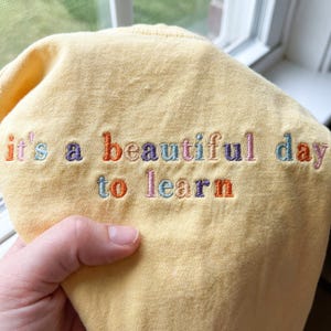 May include: A yellow sweatshirt with the embroidered phrase "it's a beautiful day to learn" in colorful thread. The text is in a playful, handwritten style, making it suitable for educational or casual wear.