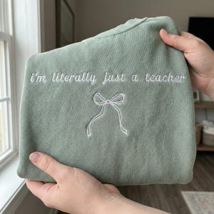 May include: Sage green sweatshirt with white embroidered text that reads "I'm literally just a teacher" and a bow design. The sweatshirt is held up by a person's hands. The sweatshirt is a crew neck style.