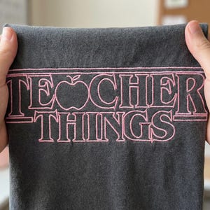 以下が含まれることがあります： ピンク色の「TEACHER THINGS」の文字が刺繍された濃いグレーのTシャツ。「TEACHER」の文字の「A」の代わりにリンゴのグラフィックが使われています。Tシャツは手で持たれています。