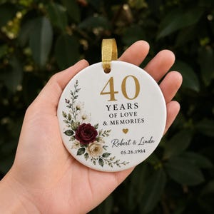 Puede incluir: Un adorno redondo de cerámica blanca con una cinta dorada. El diseño incluye un arreglo floral de rosas rojo oscuro y crema, el texto 40 YEARS OF LOVE & MEMORIES, un pequeño corazón dorado y los nombres Robert & Linda junto a 05.26.1984.