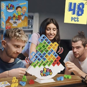 May include: A group of people playing a colorful Tetra Tower balancing game. The game features a white base with a colorful tower of blocks. The box shows the game name and cartoon illustrations. A yellow sticky note shows "48 PCS".