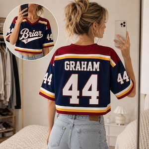 Puede incluir: Una camiseta corta de color azul marino, granate y amarillo con el nombre GRAHAM y el número 44 en blanco en la espalda. La vista frontal muestra el texto Briar U en blanco. La prenda de manga corta tiene cuello en V y rayas horizontales.