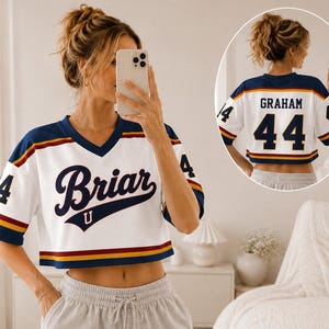 May include: A white cropped V-neck jersey with navy blue, red, and yellow stripes on the sleeves and hem. The front features the text Briar U in navy blue. The back displays the name Graham and the number 44 in navy blue with matching sleeve numbers.