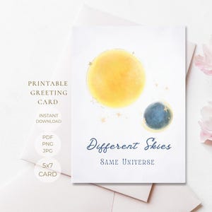 Puede incluir: Una tarjeta de felicitación blanca con un gran sol amarillo en acuarela y un planeta azul más pequeño rodeado de diminutas estrellas doradas. El texto Different Skies Same Universe aparece en letras azul oscuro y gris en la tarjeta.