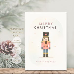 Op de afbeelding: Een witte wenskaart met een centrale illustratie van een notenkraker in roze, blauw en zwart. De tekst luidt MERRY CHRISTMAS bovenaan en Warm Holiday Wishes onderaan, omringd door kleine gouden sterren op een lichte achtergrond.
