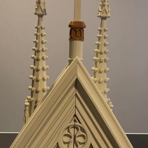 May include: A detailed architectural model in cream and brown tones. The model features a cross, decorative spires, and a roof with a carved floral design. The design is reminiscent of a church or cathedral.