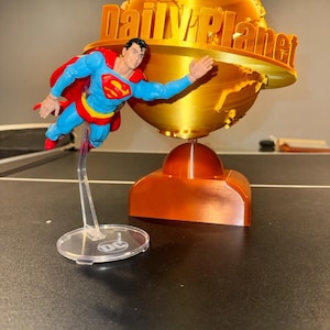 May include: A Superman action figure in flight, with a red cape and blue suit, is positioned in front of a golden Daily Planet globe. The base is orange, and the figure is supported by a clear stand with a DC logo.
