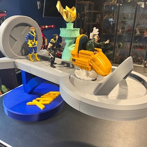 Könnte beinhalten: Ein 3D-gedrucktes X-Men-Spielset mit Cyclops, Wolverine und Professor X in seinem Hoverchair. Das Set enthält eine blaue Basis mit gelbem X-Men-Logo, eine graue Plattform und eine Nachbildung der Freiheitsstatue.