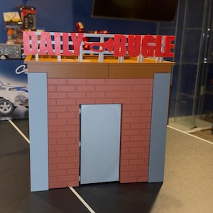 May include: A miniature model of the Daily Bugle building, featuring a red brick facade, a gray door, and the words "DAILY BUGLE" in red letters across the top. The model has a brown roof and gray side accents.