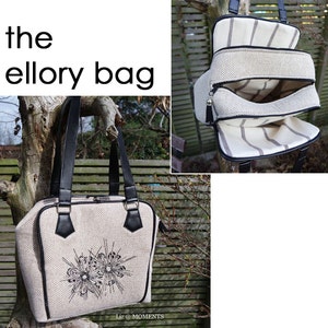 May include: A white canvas tote bag with black trim and a zip closure. The bag has a floral design embroidered on the front and the text "Liz @ MOMENTS" printed on the bottom.