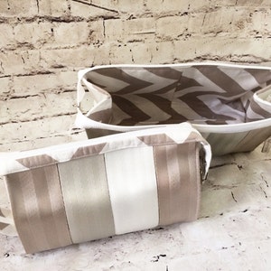 May include: Two beige and white striped cosmetic bags. One is rectangular with a handle, and the other is a larger, open-top version. Both have a chevron patterned lining.