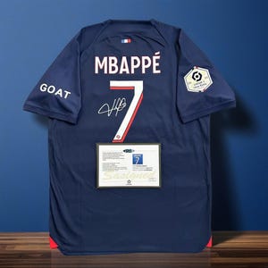 Pode incluir: Camisola de futebol azul marinho com o nome "MBAPPÉ" e o número "7" em branco. A camisola tem uma assinatura e a palavra "GOAT" na manga. Um certificado de autenticidade é exibido abaixo do número.