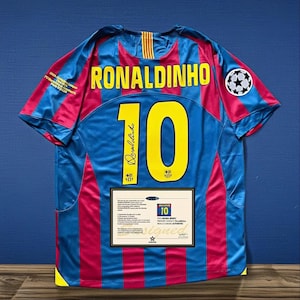 Może przedstawiać: Niebiesko-czerwona koszulka piłkarska z nazwiskiem "RONALDINHO" i numerem "10" w kolorze żółtym. Koszulka ma grafikę piłki nożnej i certyfikat autentyczności.