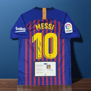 Puede incluir: Una camiseta de fútbol azul y granate del Barcelona con el nombre "MESSI" y el número "10" en amarillo en la espalda. La camiseta tiene el logo "beko" en la manga izquierda y el logo "La Liga" en la manga derecha. Un certificado de autenticidad enmarcado está debajo.
