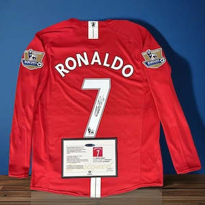 Puede incluir: Una camiseta de fútbol roja de manga larga con el nombre "RONALDO" y el número "7" en la espalda. La camiseta tiene una raya blanca en la espalda y el logo de la Premier League Barclays. Se muestra un certificado enmarcado debajo.