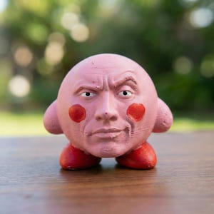 Puede incluir: Una escultura rosa de un personaje con rostro humano, mejillas rojas y pies rojos. El rostro tiene una expresión severa. La escultura está sobre una superficie de madera con un fondo verde borroso.