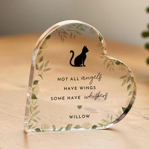 May include: Heart-shaped clear acrylic keepsake with a black cat silhouette and the text "NOT ALL angels HAVE WINGS SOME HAVE whiskers WILLOW". The edges are decorated with green and white leaf accents. A heartfelt memorial gift.