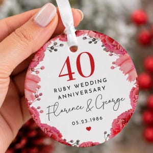 Op de afbeelding: Een rond wit keramieken ornament met het rode getal 40, de tekst Ruby Wedding Anniversary, de namen Florence & George en de datum 23-05-1986. De rand is versierd met roze en rode bloemen en een klein rood hartje.