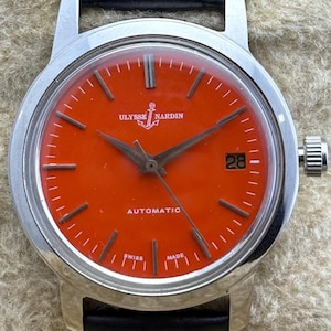 May include: A Ulysse Nardin automatic wristwatch with a vibrant orange dial and a black leather strap. The watch features a silver-toned case, a date display at the 3 o'clock position, and the words "SWISS MADE" at the bottom.