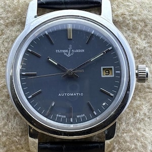 May include: A Ulysse Nardin automatic wristwatch with a black dial and a date window at the 3 o'clock position. The watch has a silver case and a black leather strap with white stitching. The watch face displays the brand name and the word "AUTOMATIC".