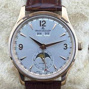 May include: A gold-cased wristwatch with a light grey face and a brown leather strap. The watch face displays the date, month, and a moon phase indicator. The brand name "JAEGER-LECOULTRE" is visible. The watch has a classic design.