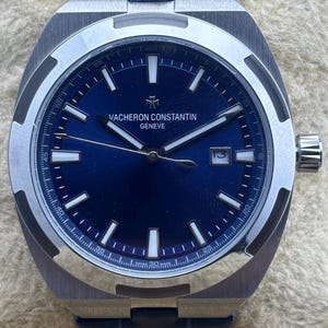 Pode incluir: Um relógio de pulso de luxo com mostrador azul e moldura prateada. O relógio apresenta o texto "VACHERON CONSTANTIN GENEVE" e um visor de data. Possui uma pulseira de couro azul.