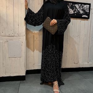 Peut inclure: Une abaya noire longue jusqu'aux pieds avec des manches longues et larges, ornée au niveau des manches et de l'ourlet d'une broderie argentée en forme de branches et de petites paillettes. Elle est portée avec une pochette monogrammée et des sandales.