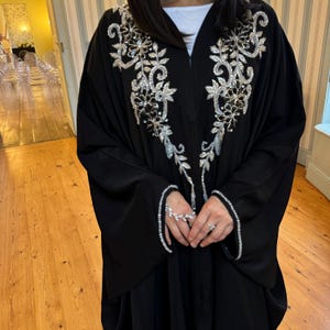 May include: A black abaya featuring detailed silver and clear sequin floral embroidery on the front and sleeve cuffs. The abaya has a flowing design and is worn over a white top. The sleeves are long and wide.