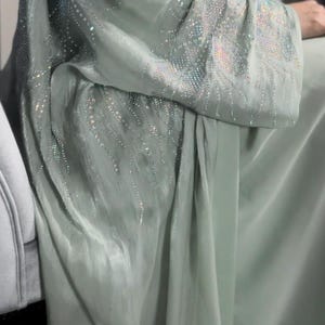 Może przedstawiać: Zwiewna, jasnozielona sukienka z misternymi kryształowymi zdobieniami na rękawach i gorsecie. Materiał układa się elegancko, tworząc miękki, mieniący się efekt. Sukienka wydaje się być wykonana z lekkiego, jedwabistego materiału.