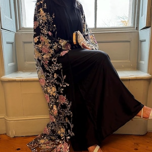 May include: A black floor-length abaya featuring long, sheer sleeves adorned with intricate floral embroidery in shades of pink, gold, and silver, accented with sparkling sequins. The garment is paired with a matching black clutch with gold trim.