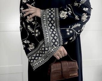 May include: A black garment with ornate, gold-colored floral embroidery on the sleeves. A small, brown, rectangular handbag with a top handle is held in the hand. The handbag has a crocodile skin texture and the word "JACQUEMUS" is printed on the front.