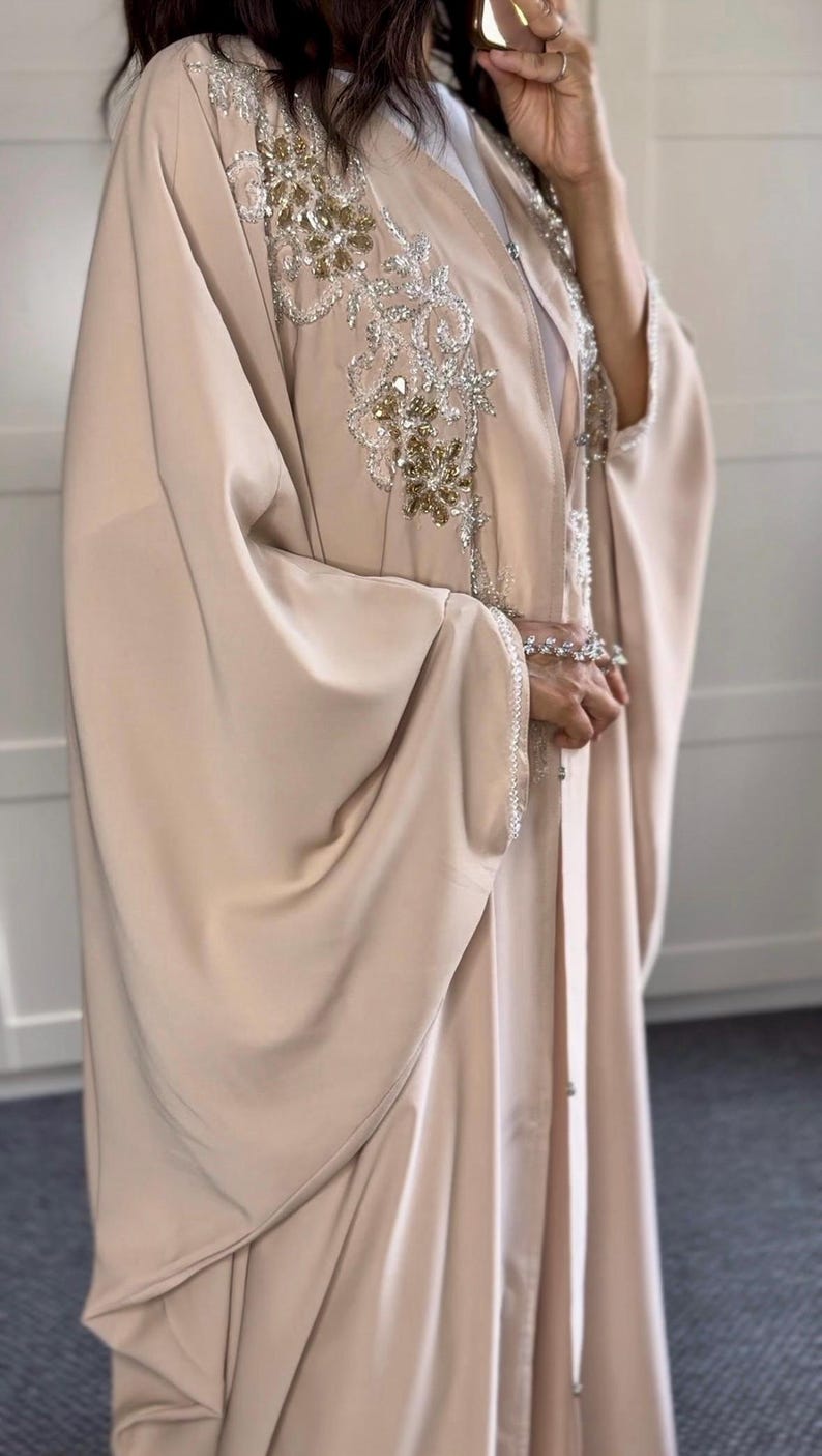 May include: A light beige abaya with detailed silver and gold embroidery on the shoulders and neckline. The sleeves are wide and the fabric drapes elegantly. The garment is buttoned down the front.
