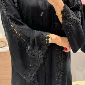 以下が含まれることがあります： 袖と前面に複雑なフローラルレースのディテールが施された黒いアバヤ。袖はフレアで、生地は微妙な光沢があります。小さな輝く装飾が衣服全体に散りばめられており、エレガントさを添えています。