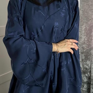 以下が含まれることがあります： 全体に同系色の蔦と葉の刺繍が施された、ダークネイビーブルーの長袖チュニック。丸首で、肩からゆったりと落ちる幅広の袖が特徴で、流れるようなシルエットを作り出しています。